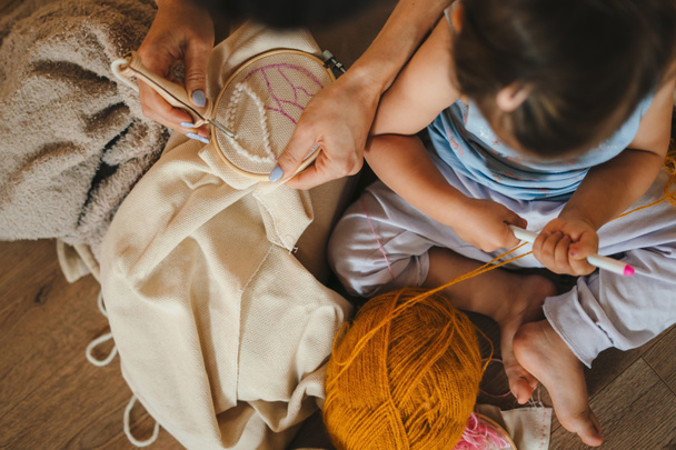 Top view of a mother and little baby daughter sitting on the floor in living room knitting with needles together. Family activity at home. People lifestyle - Foto, Bild