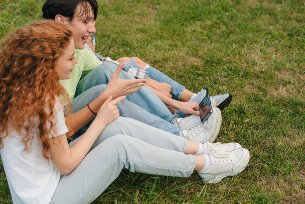 Carefree χαρούμενους φίλους καταγραφή βίντεο blog κοιτάζοντας οθόνη smartphone, κάθεται στο πράσινο γρασίδι στο πάρκο εξωτερική. Πορτρέτο για σχεδιασμό τρόπου ζωής. Κινητό τηλέφωνο - Φωτογραφία, εικόνα