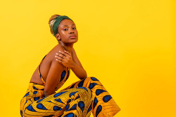 A woman of black ethnicity with traditional dress on a yellow background, sitting, commercial photography - Foto, afbeelding