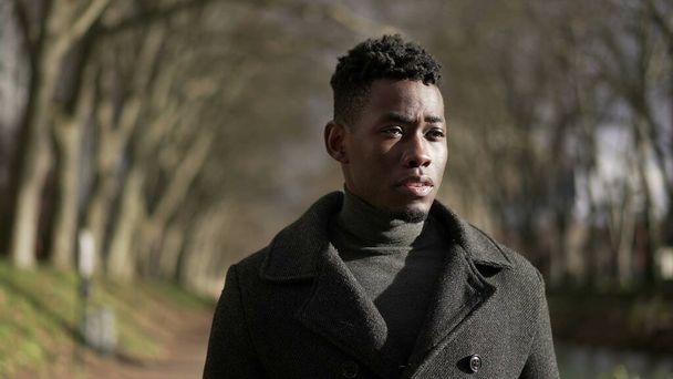 Pensive young African man walking outside in city park nature during winter season - 写真・画像