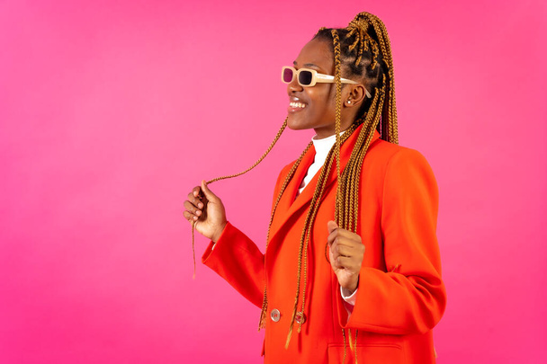 African young woman with braids on a pink background, portrait in the studio in a red suit and sunglasses - Foto, imagen