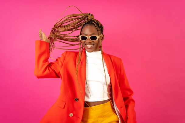 African young woman with braids on a pink background, portrait in a red suit and yellow pants smiling - Fotó, kép