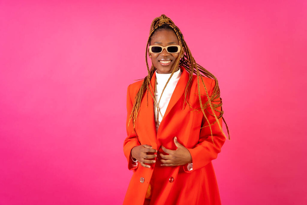 Black ethnic woman with braids on a pink background, portrait in a red suit and yellow pants smiling - Photo, Image