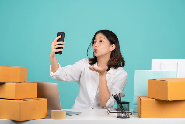 Happy Asian businesswoman sitting at her desk with laptop and brown cardboard isolated on pastel green background. She is taking a selfie with her smartphone. - Photo, Image