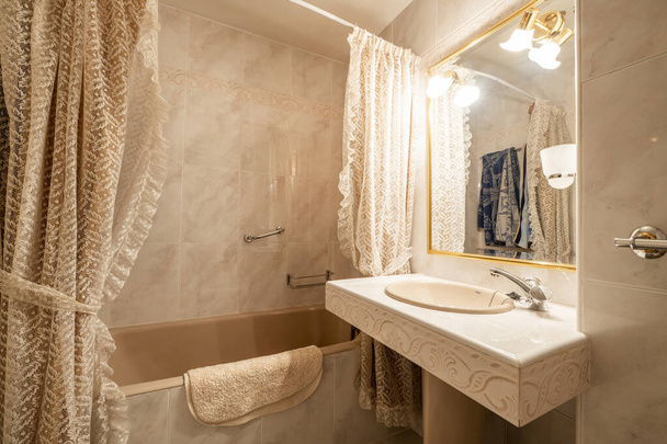Bathroom with gold-framed mirrors on the walls covered in marble-effect stoneware, cream marble floors with matching countertop with porcelain base and bathtub with rococo-style curtains - Photo, Image