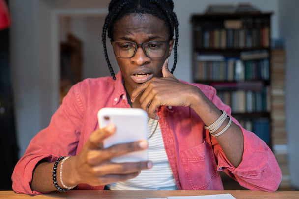 Przerażony, niespokojny Afroamerykanin czytający wiadomości w telefonie komórkowym albo porzucający studia. Niezadowolony czarny facet w okularach i luźnych ubraniach siedzi przy stole w domu ze smartfonem w ręku - Zdjęcie, obraz