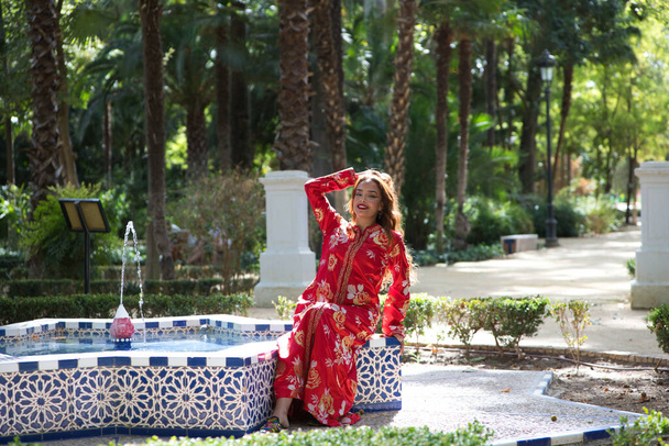 Μια όμορφη μαροκινή γυναίκα φοράει ένα παραδοσιακό μαροκινό κόκκινο φόρεμα με χρυσά και ασημένια κεντήματα. Η γυναίκα κάθεται σε ένα σιντριβάνι σε ένα πάρκο στο Seville, ενώ απολαμβάνει το ταξίδι της. - Φωτογραφία, εικόνα