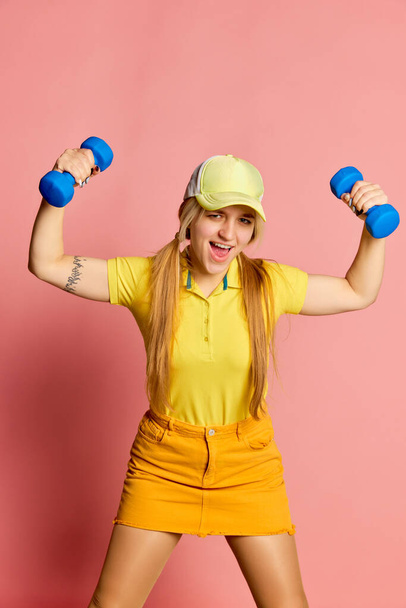 Big energy. Fitness girl. Young emotional blonde girl has fun doing exercises with dumbbells isolated on pink background. Vivid emotions, happiness, hobbies and joy. Looks delighted, excited - Foto, Imagem