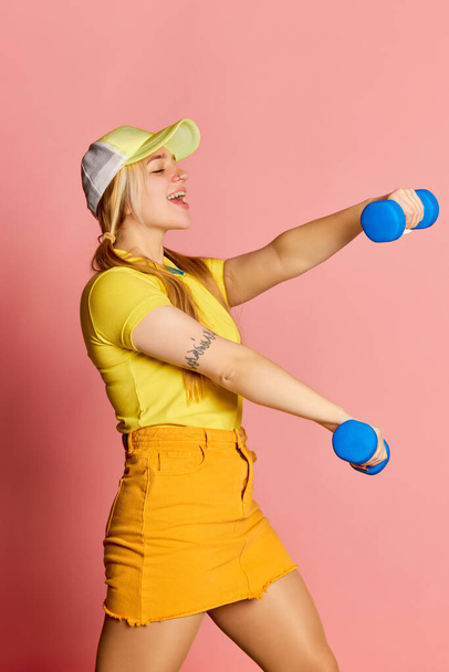 Healthy lifestyle. Fitness girl. Young emotional blonde girl has fun doing exercises with dumbbells isolated on pink background. Vivid emotions, happiness, hobbies and joy. Looks delighted, excited - Foto, Imagem