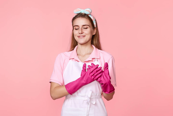 pretty friendly grateful woman in rubber gloves with hands on chest with closed eyes on pink background - Fotografie, Obrázek