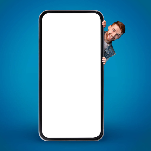 Positive Young Guy Peeking Out Of Behind Big Blank Smartphone, Happy Excited Man Showing Free Copy Space For Mobile App Or Website Design While Posing Isolated On Blue Background, Mockup - Zdjęcie, obraz