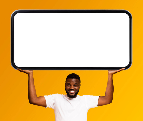 Όμορφος χαμογελαστός μαύρος άνδρας που κρατά μεγάλο κενό Smartphone πάνω από το κεφάλι, χαρούμενος αφρικανός Αμερικανός τύπος που επιδεικνύει το κινητό τηλέφωνο με τη λευκή οθόνη ενώ στέκεται απομονωμένος στο κίτρινο φόντο, Mockup - Φωτογραφία, εικόνα