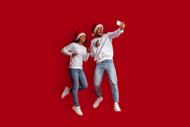 Веселе красиве афро-американське подружжя стрибає в повітря, роблячи селфі під час святкування Різдва разом, вдягаючи Санта-Капелюхи, користуючись мобільним телефоном, ізольованим на червоному студійному фоні. - Фото, зображення
