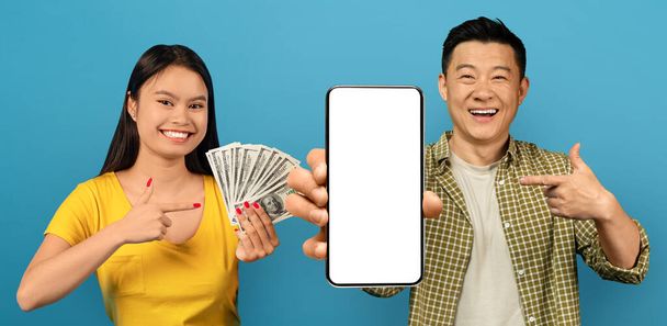 Онлайн ставки и казино, коллаж. Улыбающаяся азиатская пара, наслаждающаяся победой в синем, играющая в азартные игры онлайн, указывающая на сотовый телефон с белым пустым экраном, показывающая кучу денег, рекомендующая хорошее приложение - Фото, изображение