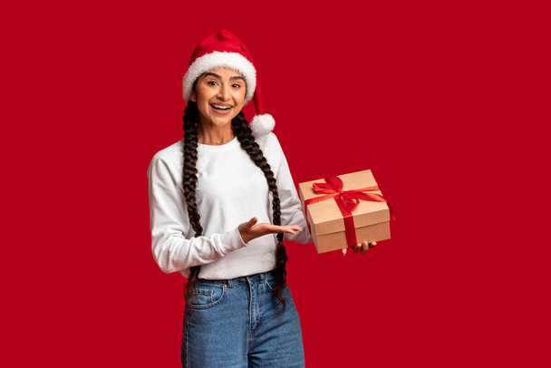 Holiday Bonus. Beautiful Arab Woman In Santa Hat Holding Wrapped Gift Box, Cheerful Young Middle Eastern Female Pointing At Present In Hand While Posing Over Red Studio Background, Copy Space - 写真・画像