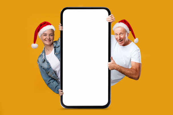 Happy european elderly woman and man in Santa Claus hats show thumb up with big phone with blank screen isolated on orange background. Offer, ad and app, recommendation for New Year and Christmas - Photo, Image