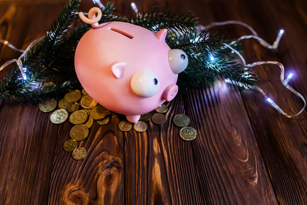 Banco porquinho em uma mesa de fazenda dupla com moedas espalhadas e guirlandas com luzes. Decoração de Natal. Gastar em férias de Ano Novo - Foto, Imagem