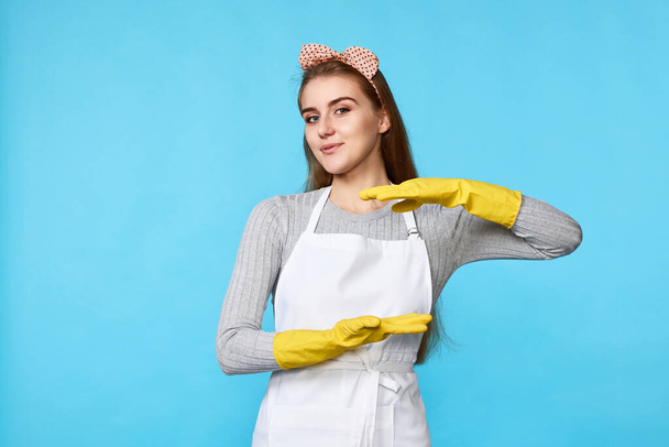 beautiful woman in yellow rubber gloves and cleaner apron showing measure symbol gesture isolated on blue background. cleaning - Fotografie, Obrázek