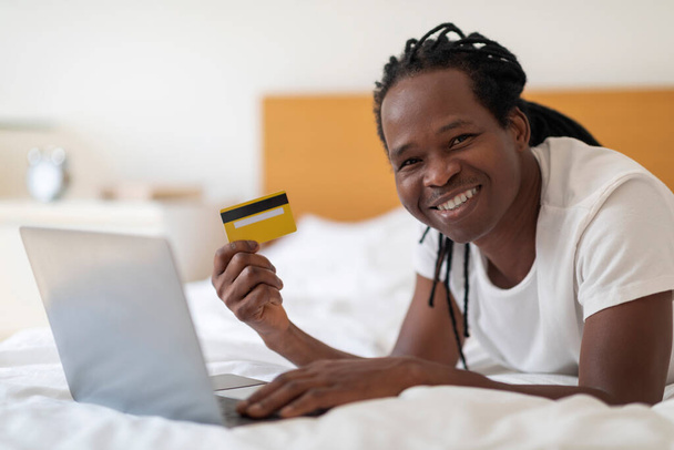Shopping en ligne. Homme noir souriant utilisant un ordinateur portable et une carte de crédit tout en se relaxant au lit à la maison, jeune homme afro-américain heureux effectuant des paiements Internet avec ordinateur tout en se reposant dans la chambre - Photo, image