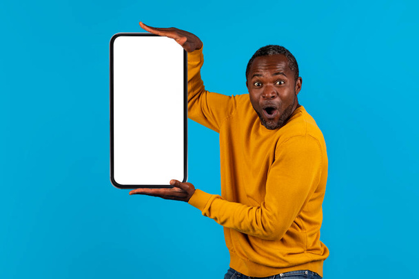 Erstaunlich emotionale schöne reife schwarze Mann hält Handy mit weißen leeren Bildschirm in beiden Händen, Attrappe, isoliert auf blauem Studiohintergrund, zeigt schönes Online-Angebot - Foto, Bild