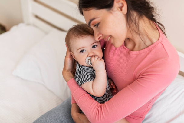 Loving mother cuddling and resting with her infant daughter on bed, mom holding cute newborn baby on hands, bonding together while relaxing at home, copy space - Photo, Image