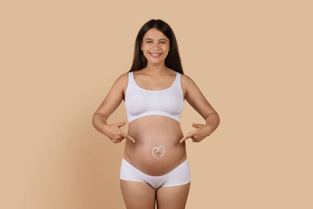 Счастливая беременная женщина в нижнем белье, указывая на форму сердца, сделанную из крема вокруг пупка, портрет веселой ожидающей женщины, наслаждающейся кожей во время беременности, бежевого фона, копирования пространства - Фото, изображение