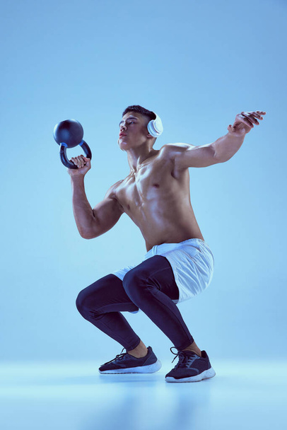Portret młodego muskularnego mężczyzny w słuchawkach treningowych, podnoszącego ciężar i robiącego przysiady odizolowane na niebieskim tle w neonowym świetle. Pojęcie sportu, sprawności, zdrowego i aktywnego stylu życia, motywacji - Zdjęcie, obraz