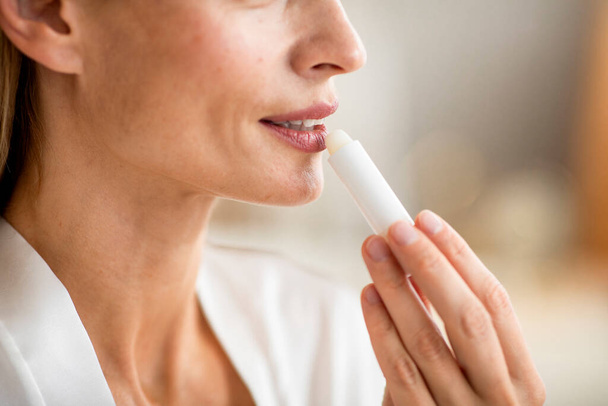 Beauty treatments concept. Middle aged woman applying balm on her lips, taking care and doing morning routine in bathroom interior, closeup, cropped - Photo, Image