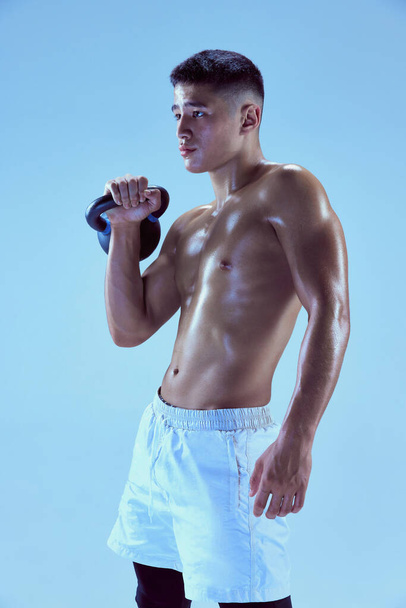 Portrait of young muscular man training, posing with weight isolated over blue background in neon light. Sportive body. Concept of sport, fitness, healthy and active lifestyle, motivation, workout - Photo, Image