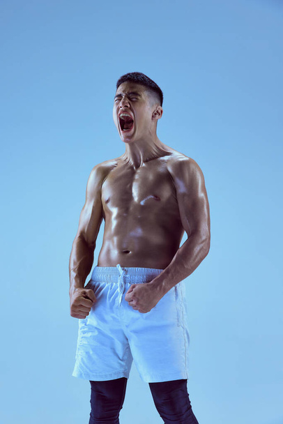 Portrait of young muscular man training, shouting to rise sportive spirit isolated over blue background in neon light. Concept of sport, fitness, healthy and active lifestyle, motivation, workout - Photo, Image