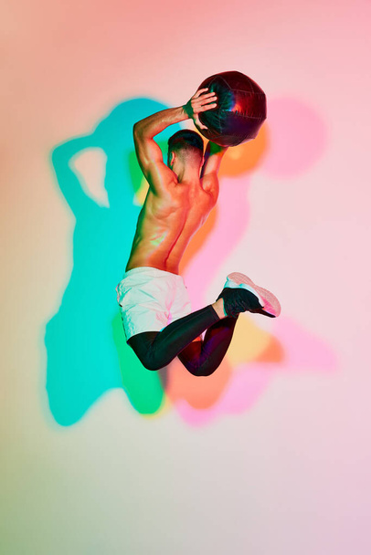 Young muscular man training with med ball in a jump isolated over pink background in neon light. Multicolored shadow. Concept of sport, fitness, healthy and active lifestyle, motivation, workout - Foto, Imagem