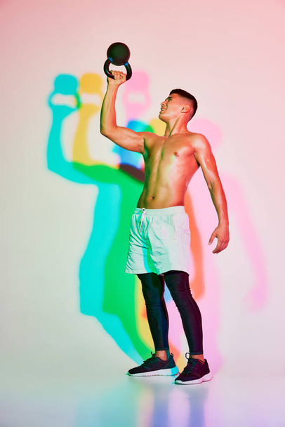 Entrenamiento de hombre musculoso joven, levantando peso aislado sobre fondo rosa en luz de neón. Sombra multicolor. Manos fuertes. Concepto de deporte, fitness, estilo de vida saludable y activo, motivación, entrenamiento - Foto, imagen