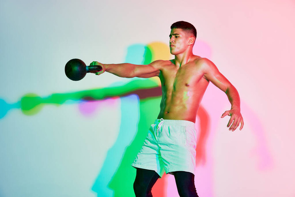 Portret van jonge gespierde man training, tillen gewicht geïsoleerd over roze achtergrond in neon licht. Meerkleurige schaduw. Concept sport, fitness, gezonde en actieve levensstijl, motivatie, training - Foto, afbeelding