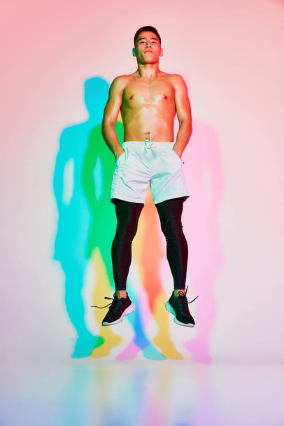 Portrait of young muscular man training, jumping isolated over pink background in neon light. Multicolored shadow. Concept of sport, fitness, healthy and active lifestyle, motivation, workout - Photo, Image