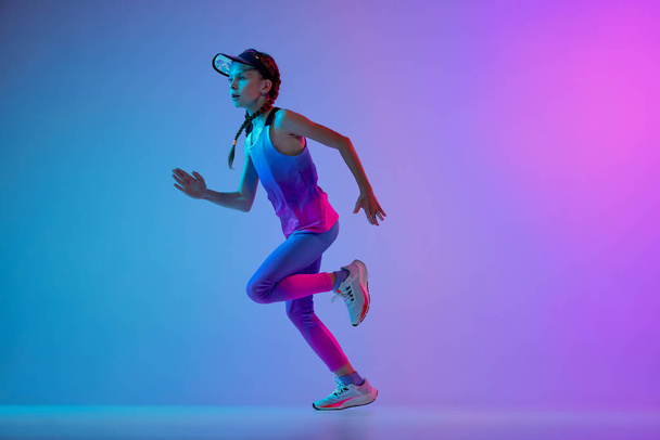 Champion. Sportive little girl, junior runner in stylish sportswear and cap running isolated on gradient pink-blue background in neon. Concept of sport, fashion, fitness and education, beauty - Foto, imagen