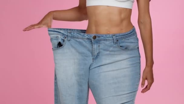 Успішна втрата ваги. Крупним планом знімок невпізнаваної стрункої жінки в джинсових штанях величезного розміру, демонструючи своє тіло, рожевий студійний фон, повільний рух, вільний простір
 - Кадри, відео