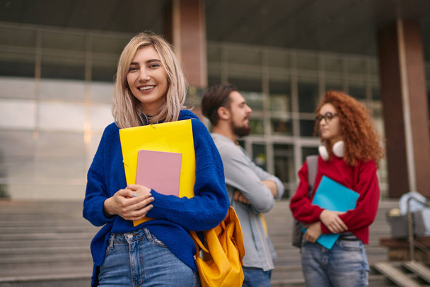 Cheerful blond woman with backpack and stationery smiling and looking at camera while standing near talking friends outside college building in daytime - 写真・画像