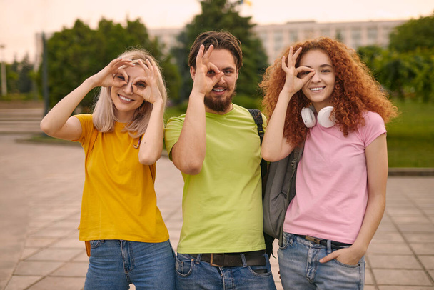 Glad man and women in colorful t shirts gesturing okay near eyes and looking at camera with smile, while spending time in park after university studies - Foto, Imagen