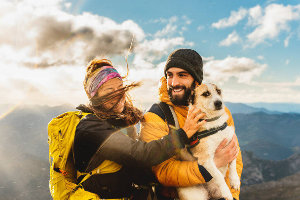 coppia di escursionisti sotto la pioggia facendo un percorso di montagna con il loro cane. Alpinista che tiene il cane tra le braccia con un paesaggio montuoso sullo sfondo. donna cane da accarezzare - Foto, immagini
