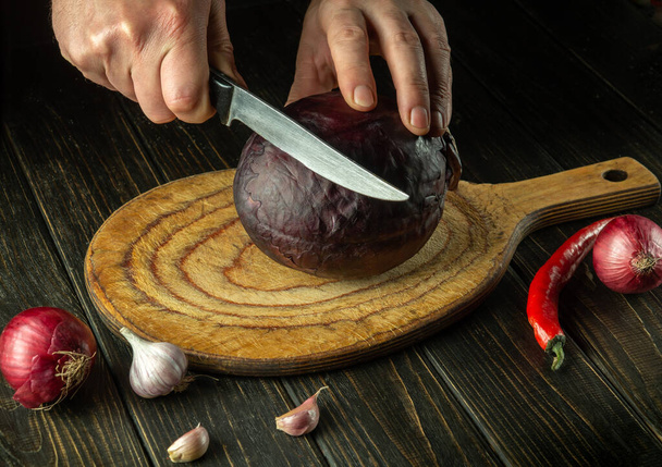 De handen van de kok snijden rode kool met een mes op een keukensnijplank. Vegetarische gerechten thuis koken. - Foto, afbeelding