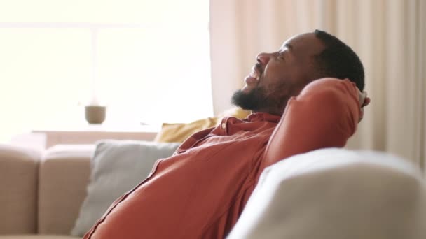 Удовлетворение. Вид сбоку портрет счастливого беззаботного африканского мужчины, отдыхающего дома на диване, опирающегося на руки и расслабляющего, замедленного движения, свободного пространства - Кадры, видео