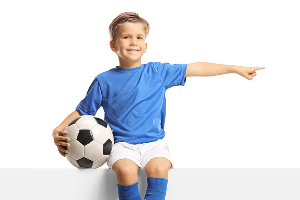 Chłopiec w niebieskiej koszulce siedzi na pustym panelu z piłką nożną i wskazuje na bok odizolowany na białym tle - Zdjęcie, obraz