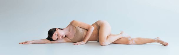 full length of barefoot and young woman with vitiligo lying in beige lingerie on grey background, banner - Zdjęcie, obraz