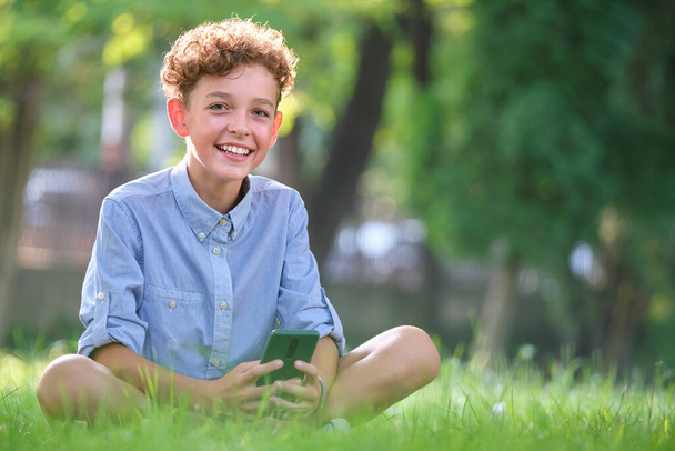 Fiatal fiú nézegeti az okostelefonját a nyári parkban. Mobiltelefon használata szabadidős és oktatási célokra. - Fotó, kép