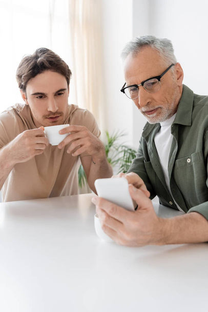 senior man in eyeglasses showing smartphone to young son drinking coffee in kitchen - Photo, Image