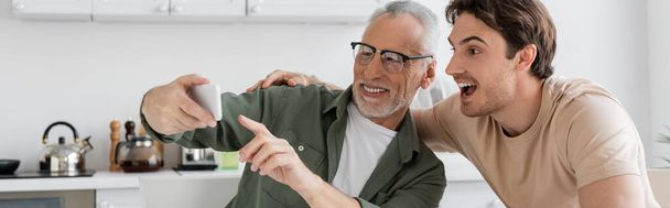 Χαρούμενος άνδρας με γυαλιά που δείχνει το smartphone κοντά έκπληκτος γιος στην κουζίνα, πανό - Φωτογραφία, εικόνα