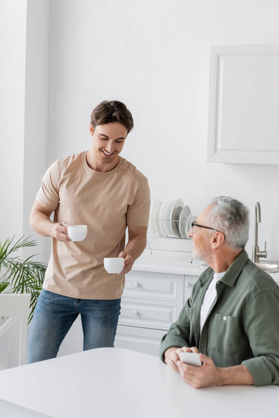 young man smiling while holding coffee cups near dad sitting with smartphone in kitchen - Photo, Image
