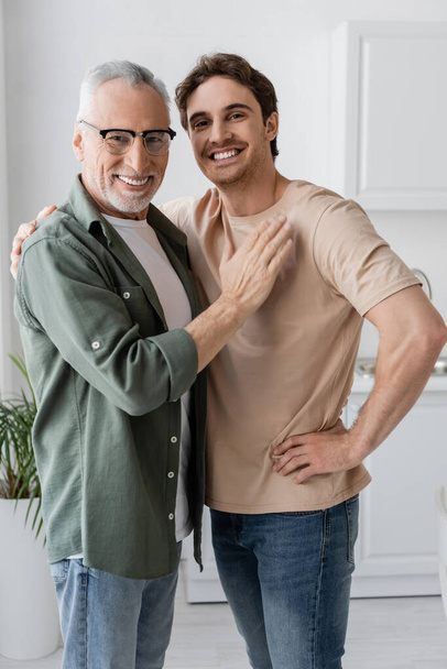 young and smiling man hugging shoulders of happy father touching his chest and smiling at camera - Photo, Image