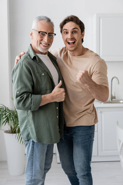 vrolijk vader en zoon tonen als en succes gebaren terwijl kijken naar de camera in de keuken - Foto, afbeelding