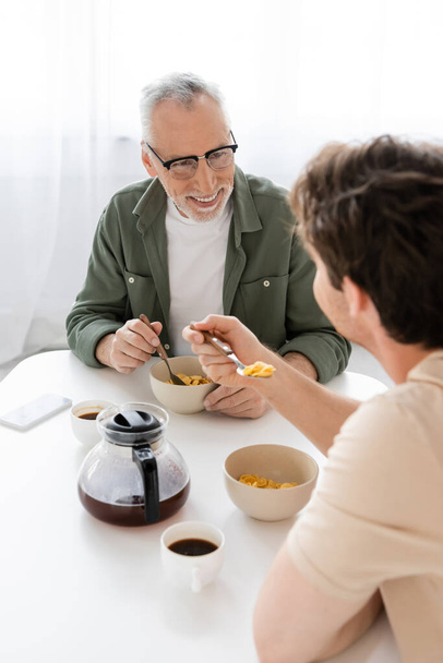 Улыбающийся мужчина в очках смотрит на маленького сына, который ест кукурузные хлопья на завтрак - Фото, изображение